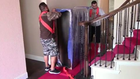 mover carrying a wrapped dresser on his back with a red lifting strap while a second mover spots from the stairs, face visible